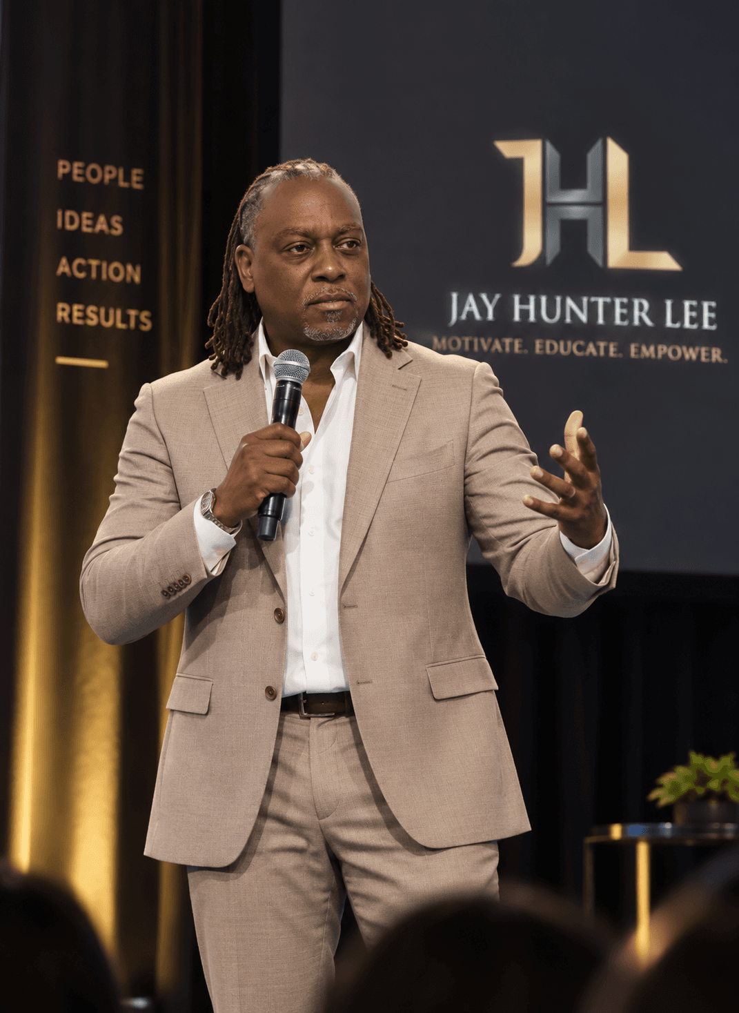 Jay Hunter Lee speaking on stage in a tan suit, holding a microphone with his JHL Motivate Educate Empower backdrop behind him